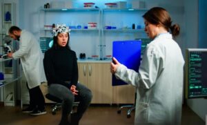 A patient wearing a neural monitoring device sits in a lab, while a healthcare professional holds a clipboard, discussing behavioral CPT codes for mental health assessment.